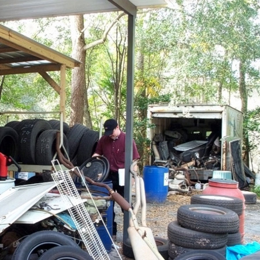 Man working among stacked tires and scrap metal in a wooded yard with a small shed filled with tools and materials.
